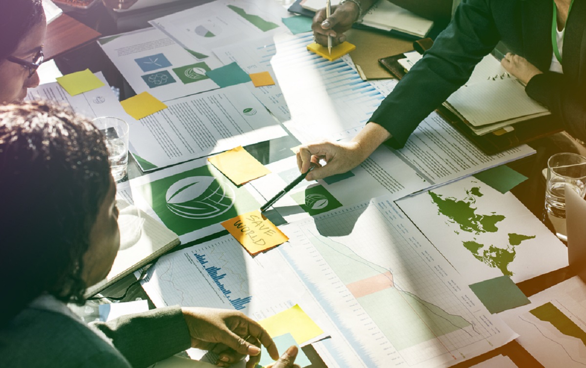 A table with maps and different documents with people pointing at a post it with 'Save world' written on it